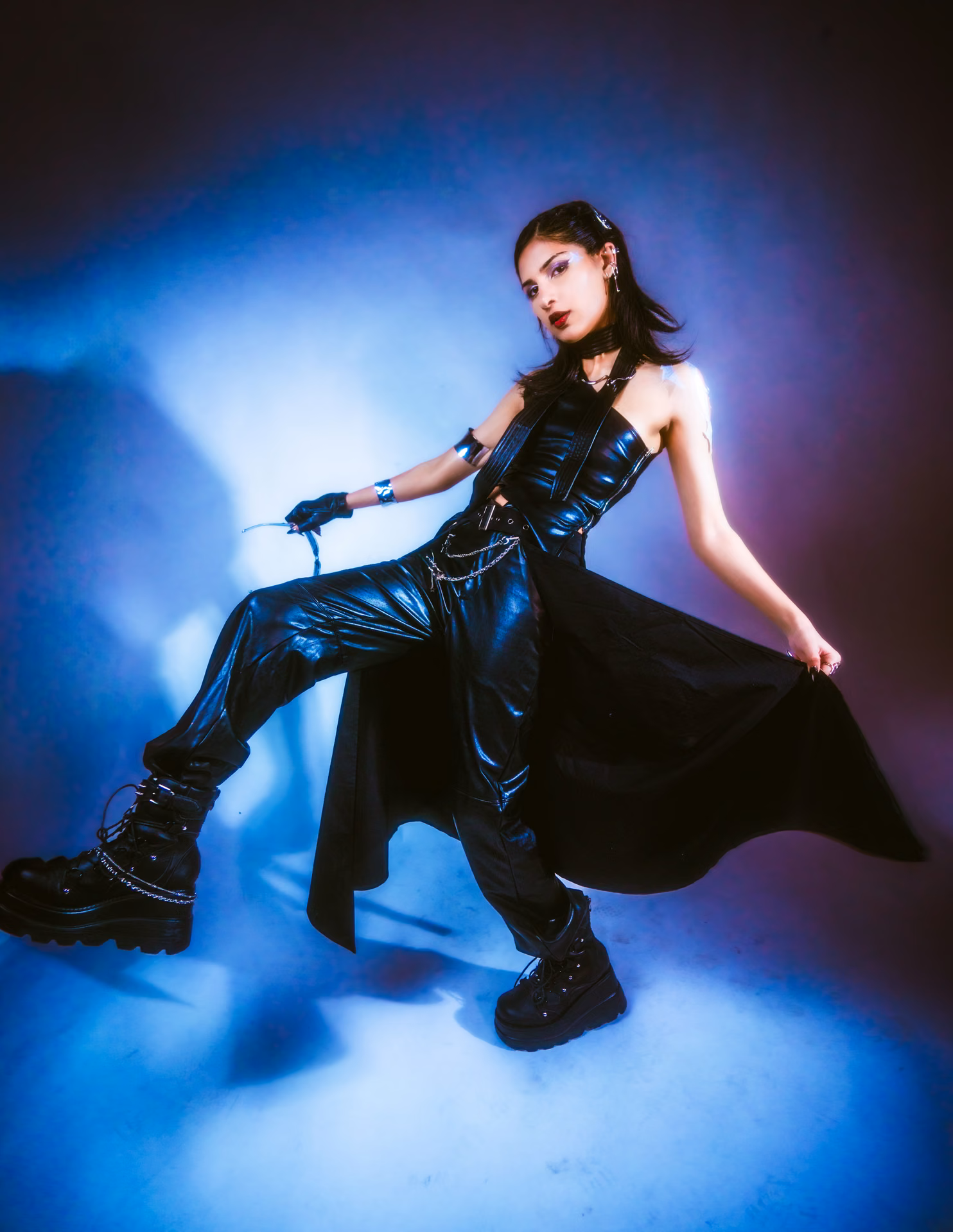 Black leather, a chain belt, and tiny chrome shades under a blue-to-magenta gel; a glam-dark character study lit for maximum edge.
