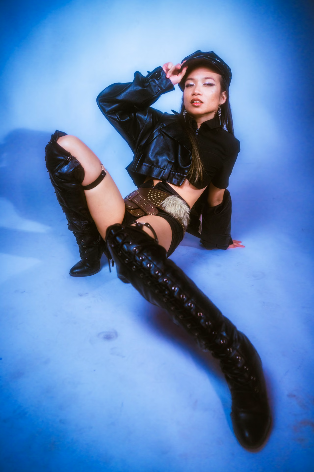 Black leather, a chain belt, and tiny chrome shades under a blue-to-magenta gel; a glam-dark character study lit for maximum edge.