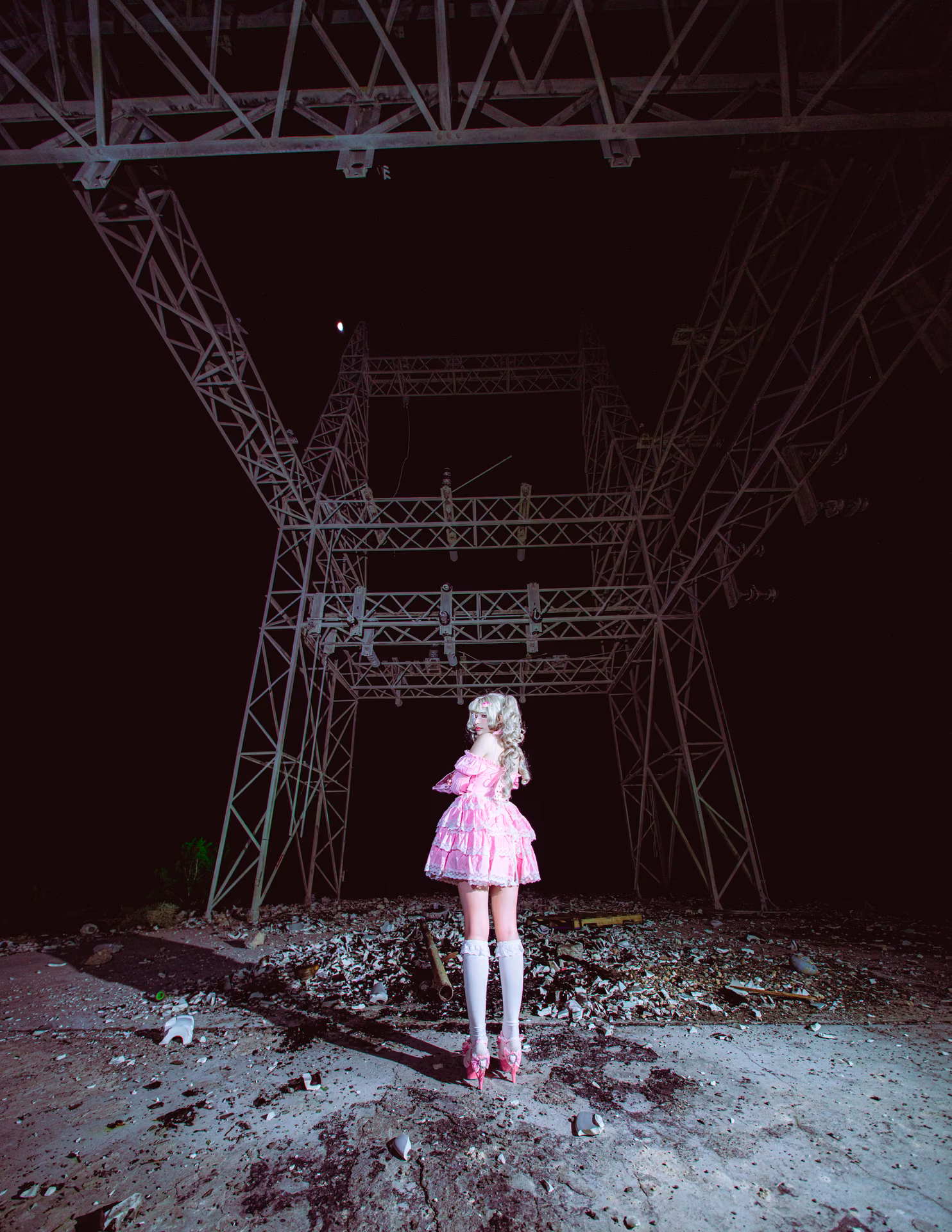 A pink frilled lolita stands alone beneath towering power pylons at night, lit like a single bloom in a steel wasteland — Davila's sweetest costume against his harshest set.