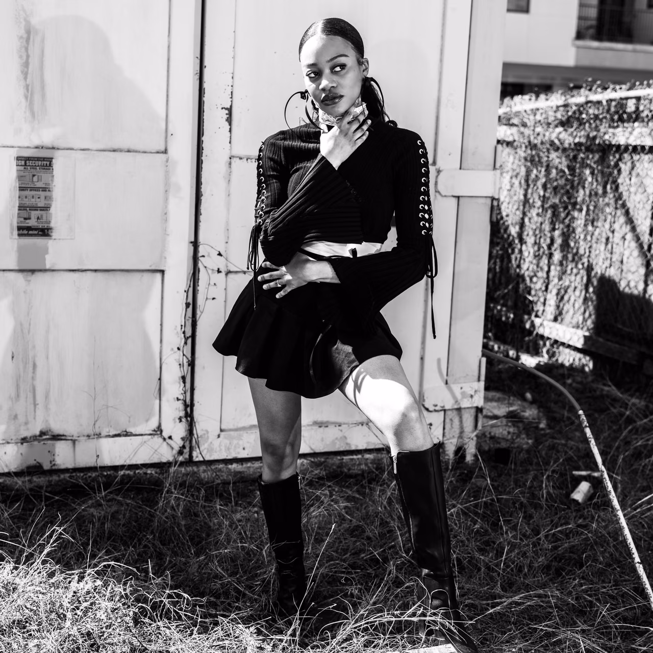 High-contrast monochrome against a corrugated shed and dry grass: lace-up sleeves, tall boots, a hand at the throat. Davila strips the color to leave only line and attitude.