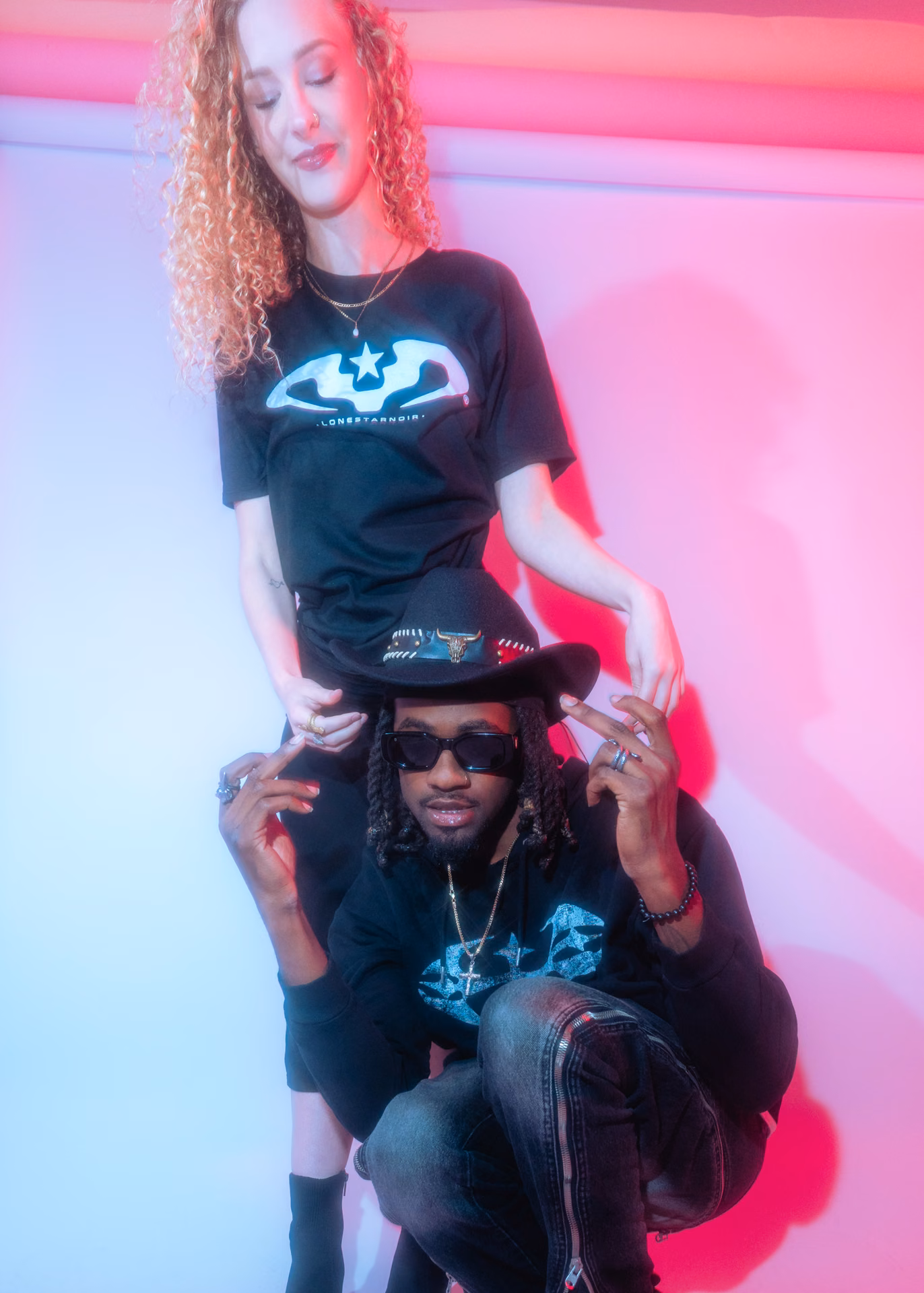 A two-up lookbook frame in saturated pink: a Lone Star Noir tee, a longhorn-banded cowboy hat, peace signs thrown into colored gel. Streetwear shot like a record cover.