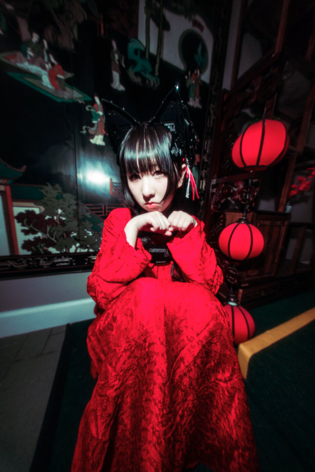 Under red lanterns on a mall staircase, a cat-eared figure in red lace cradles a branch of yellow blossom; a wide-angle frame that lets the location's color carry the mood.