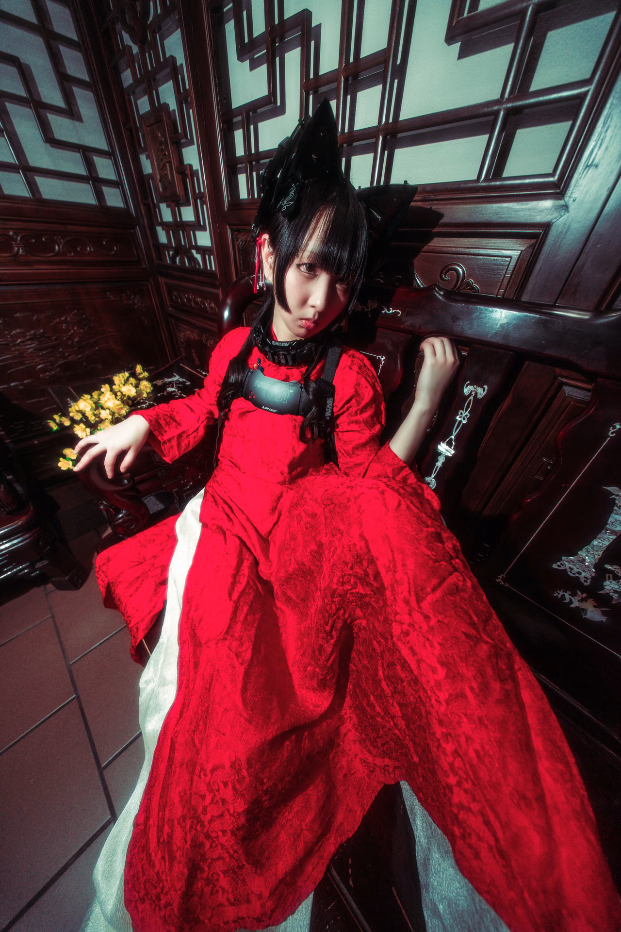 Under red lanterns on a mall staircase, a cat-eared figure in red lace cradles a branch of yellow blossom; a wide-angle frame that lets the location's color carry the mood.