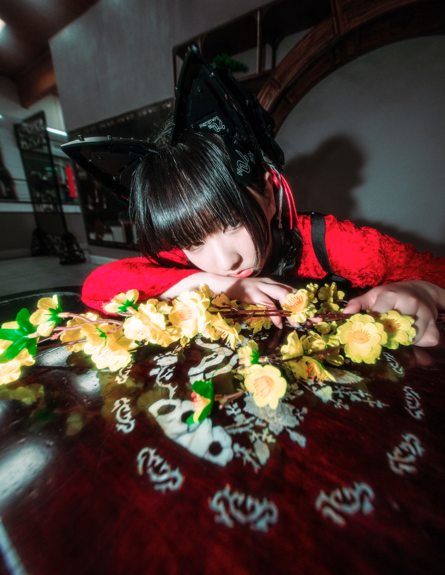 Under red lanterns on a mall staircase, a cat-eared figure in red lace cradles a branch of yellow blossom; a wide-angle frame that lets the location's color carry the mood.