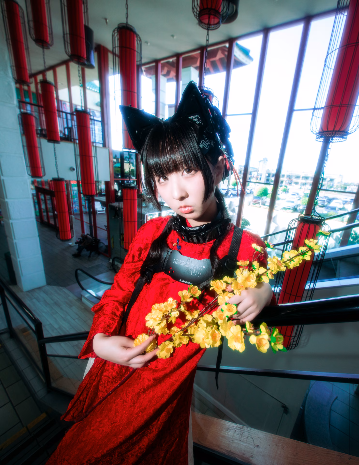 Under red lanterns on a mall staircase, a cat-eared figure in red lace cradles a branch of yellow blossom; a wide-angle frame that lets the location's color carry the mood.