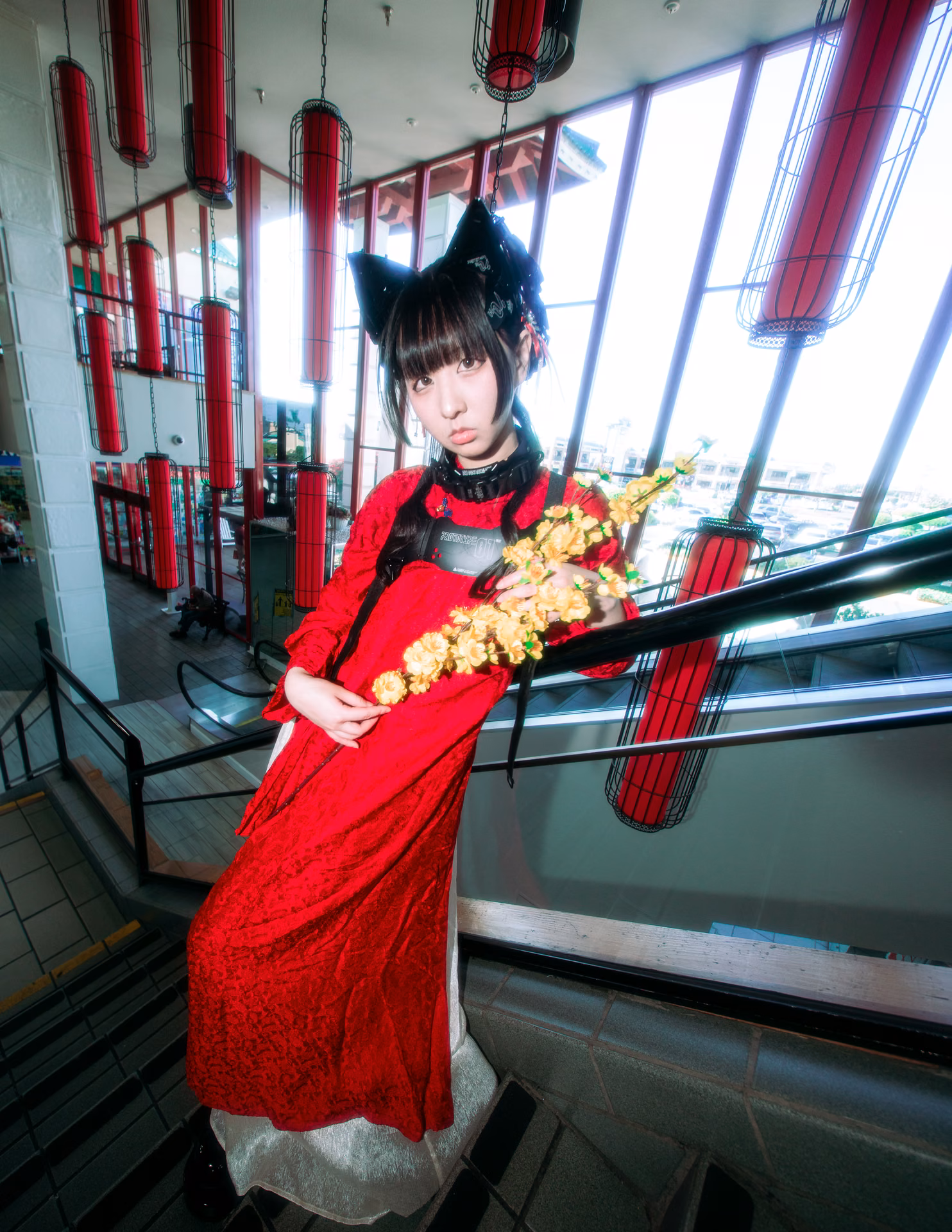 Under red lanterns on a mall staircase, a cat-eared figure in red lace cradles a branch of yellow blossom; a wide-angle frame that lets the location's color carry the mood.