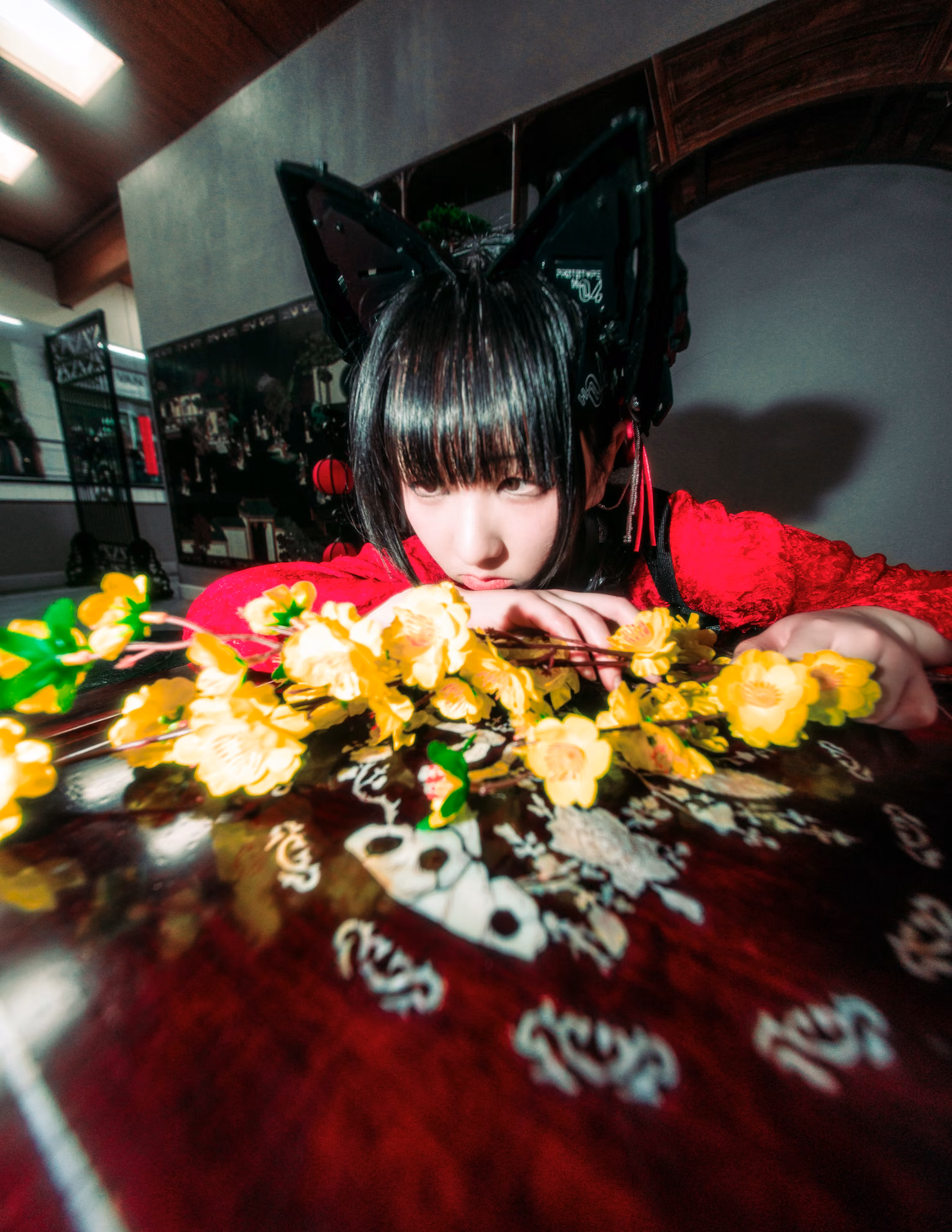 Under red lanterns on a mall staircase, a cat-eared figure in red lace cradles a branch of yellow blossom; a wide-angle frame that lets the location's color carry the mood.
