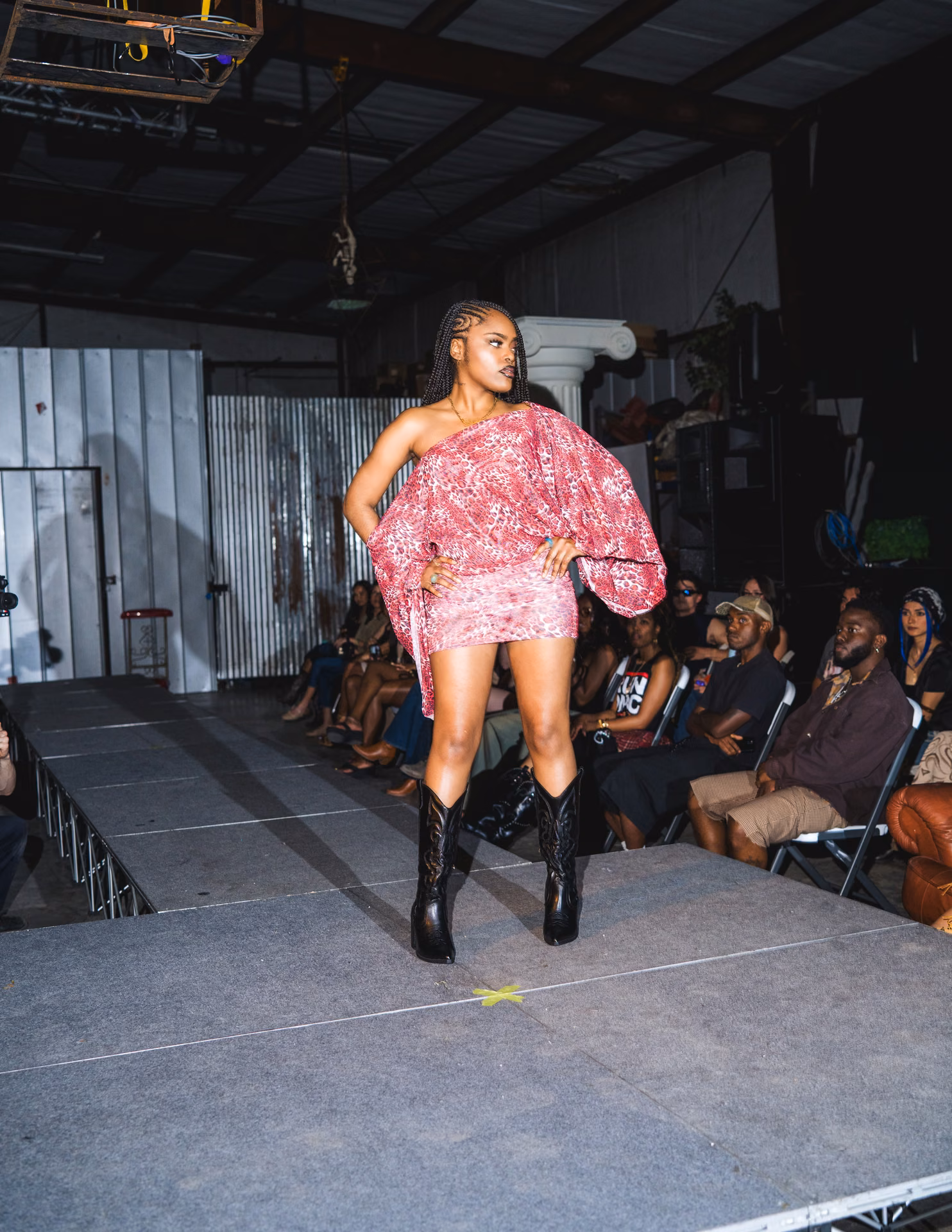 A pink off-shoulder mini and black cowboy boots cross a warehouse runway past a fallen Greek column; Texas swagger caught inside the live-show frame.