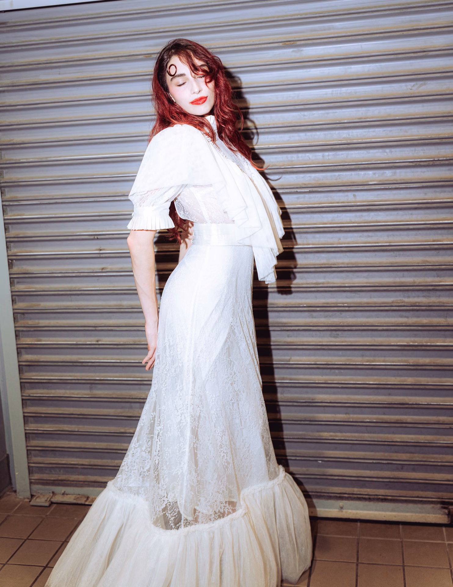 Flash hits wet auburn hair and a swirl of white lace against a metal shutter — a night-alley editorial caught mid-motion, romantic and a little feral.