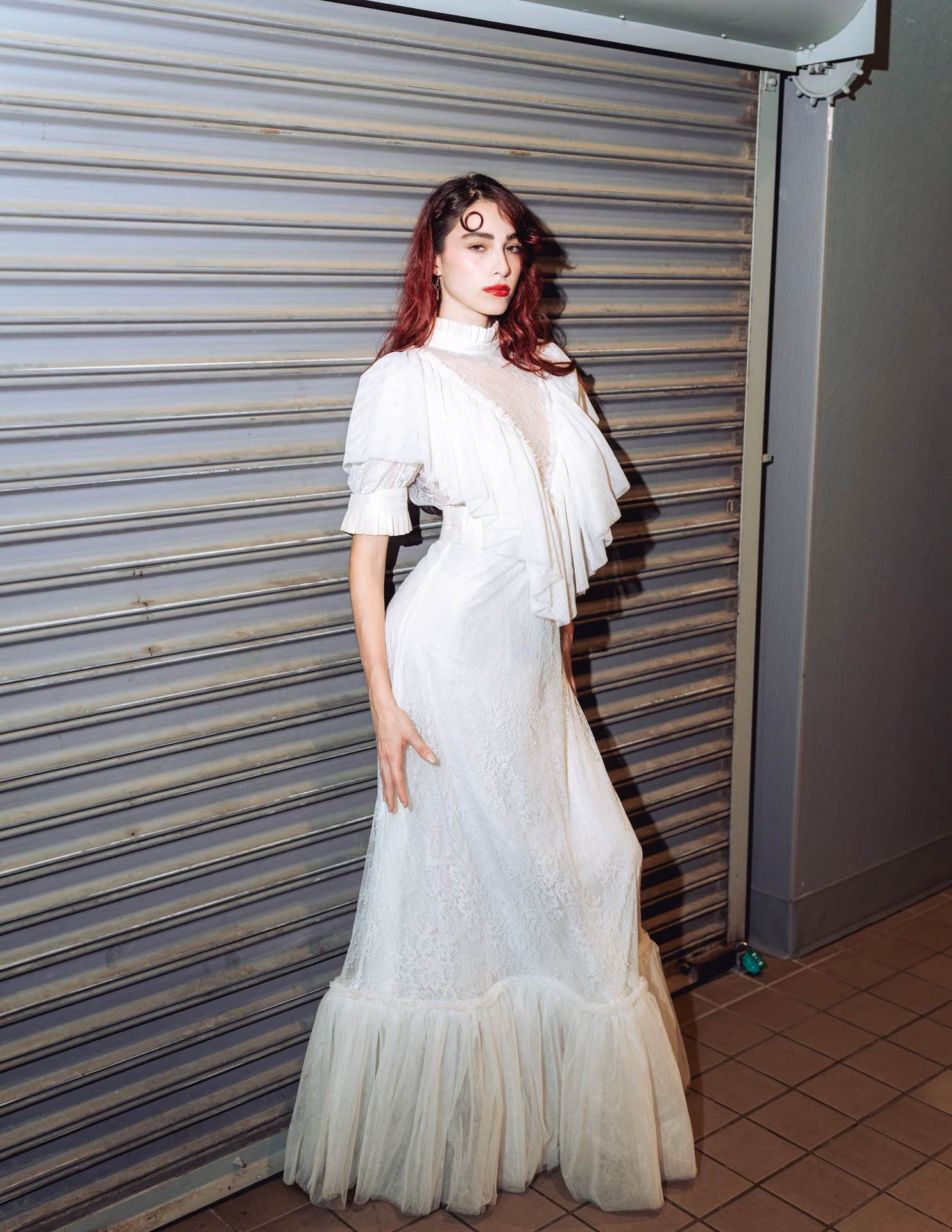 Flash hits wet auburn hair and a swirl of white lace against a metal shutter — a night-alley editorial caught mid-motion, romantic and a little feral.