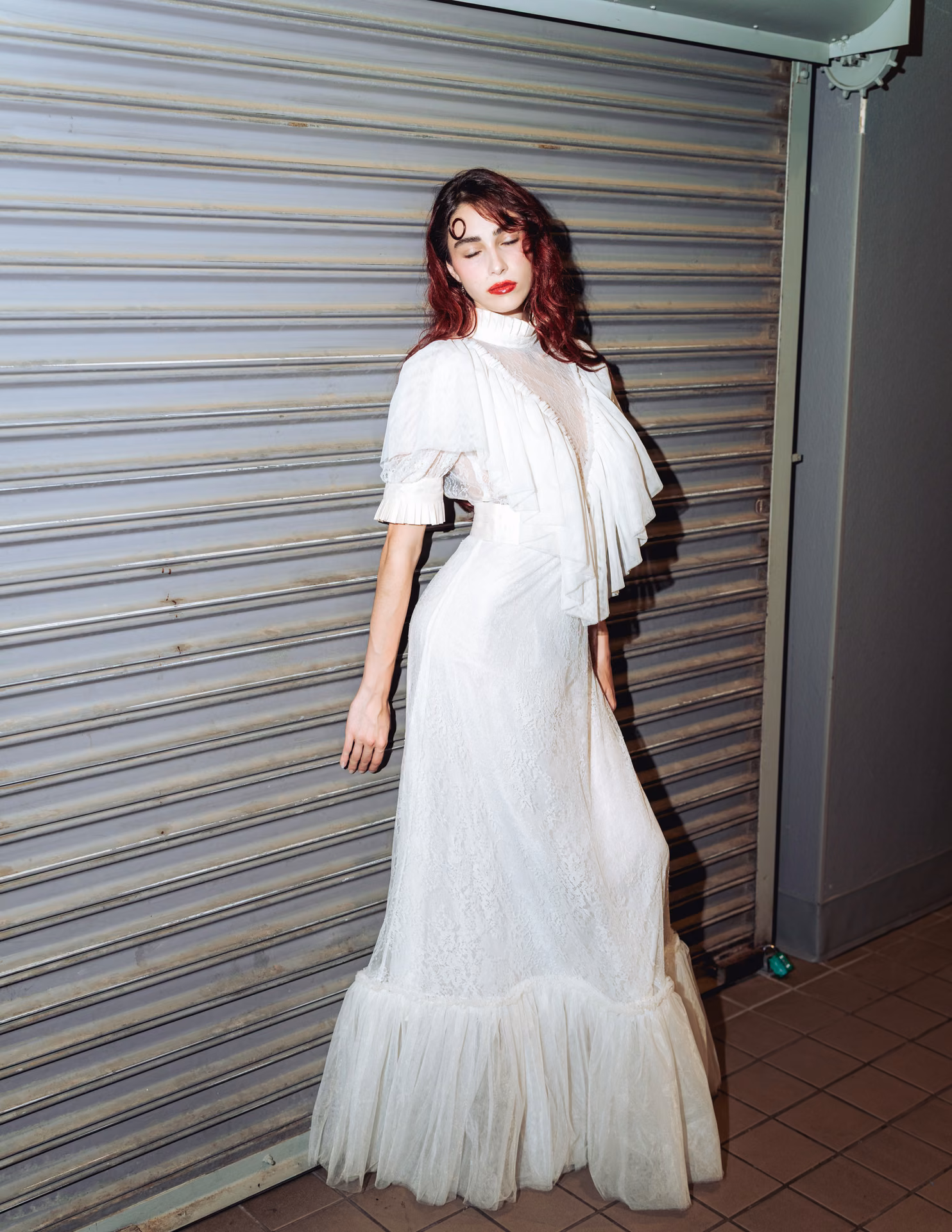 Flash hits wet auburn hair and a swirl of white lace against a metal shutter — a night-alley editorial caught mid-motion, romantic and a little feral.