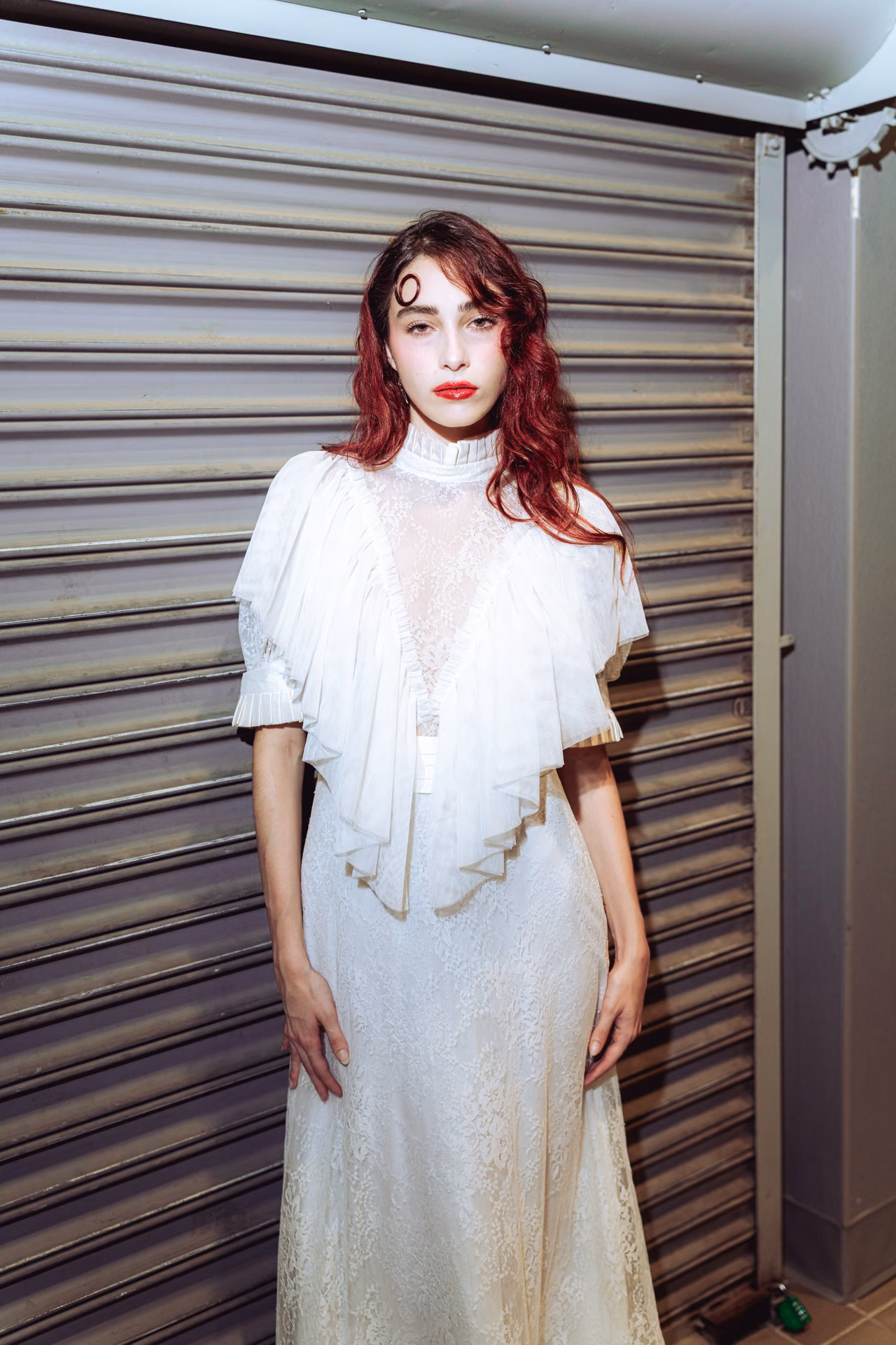 Flash hits wet auburn hair and a swirl of white lace against a metal shutter — a night-alley editorial caught mid-motion, romantic and a little feral.