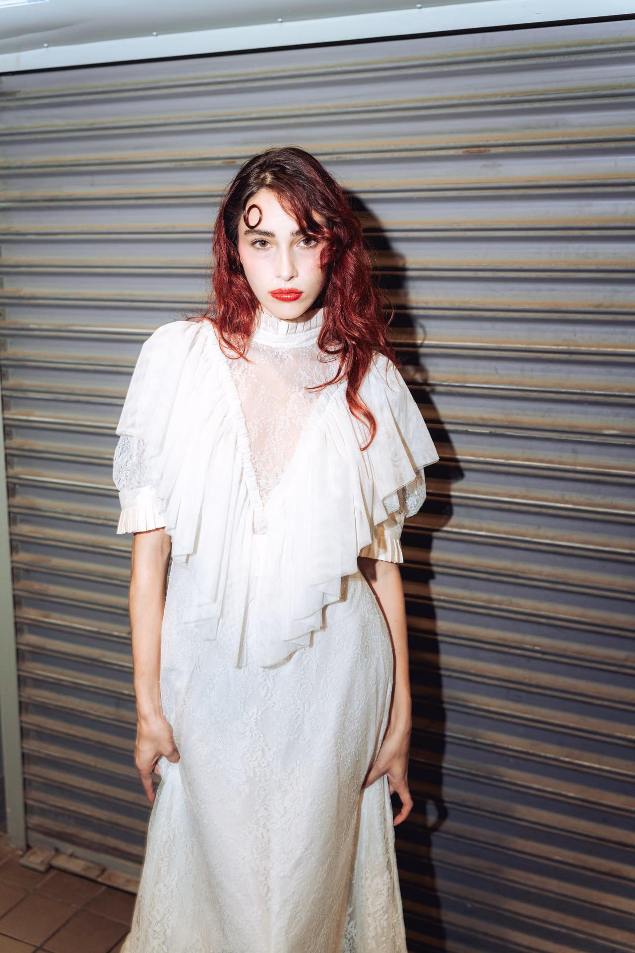Flash hits wet auburn hair and a swirl of white lace against a metal shutter — a night-alley editorial caught mid-motion, romantic and a little feral.