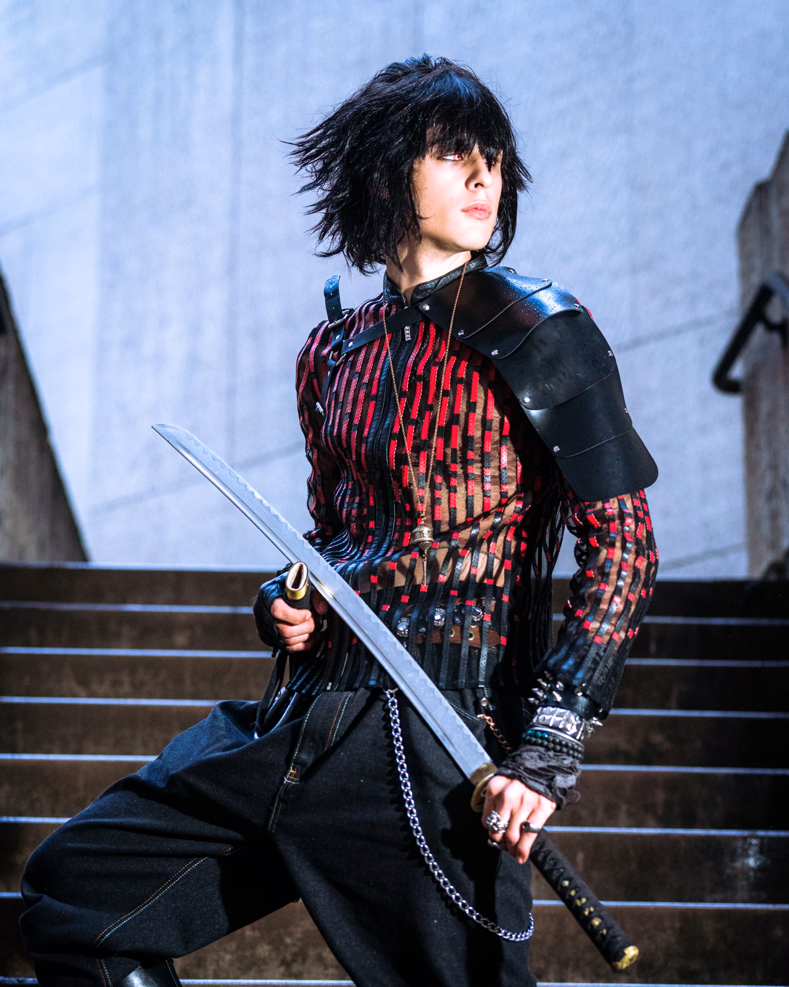 A Final Fantasy lead leans against a subway bollard at the foot of an escalator — fur pauldron, armored gauntlet, the underground light doing the cinema.