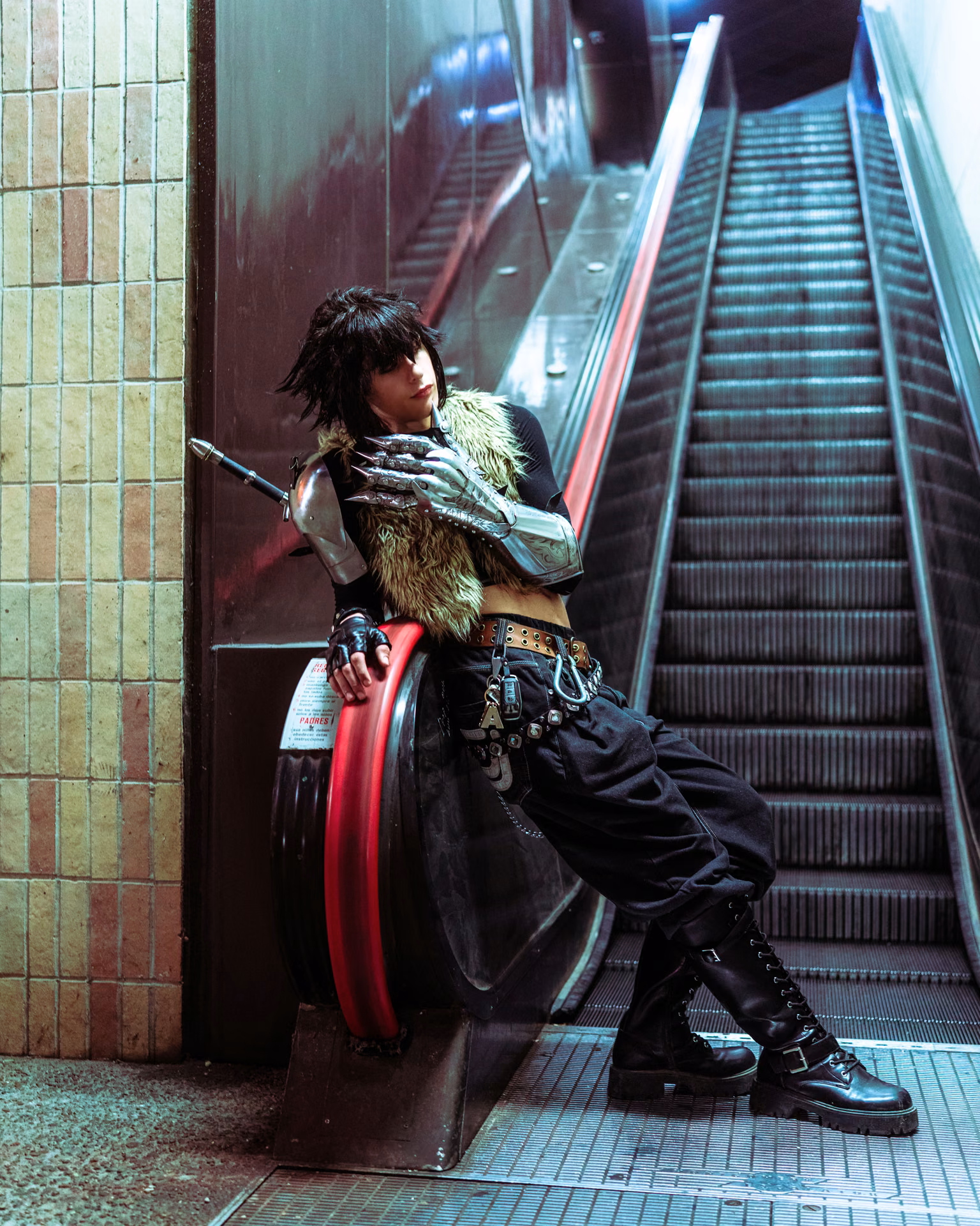 A Final Fantasy lead leans against a subway bollard at the foot of an escalator — fur pauldron, armored gauntlet, the underground light doing the cinema.