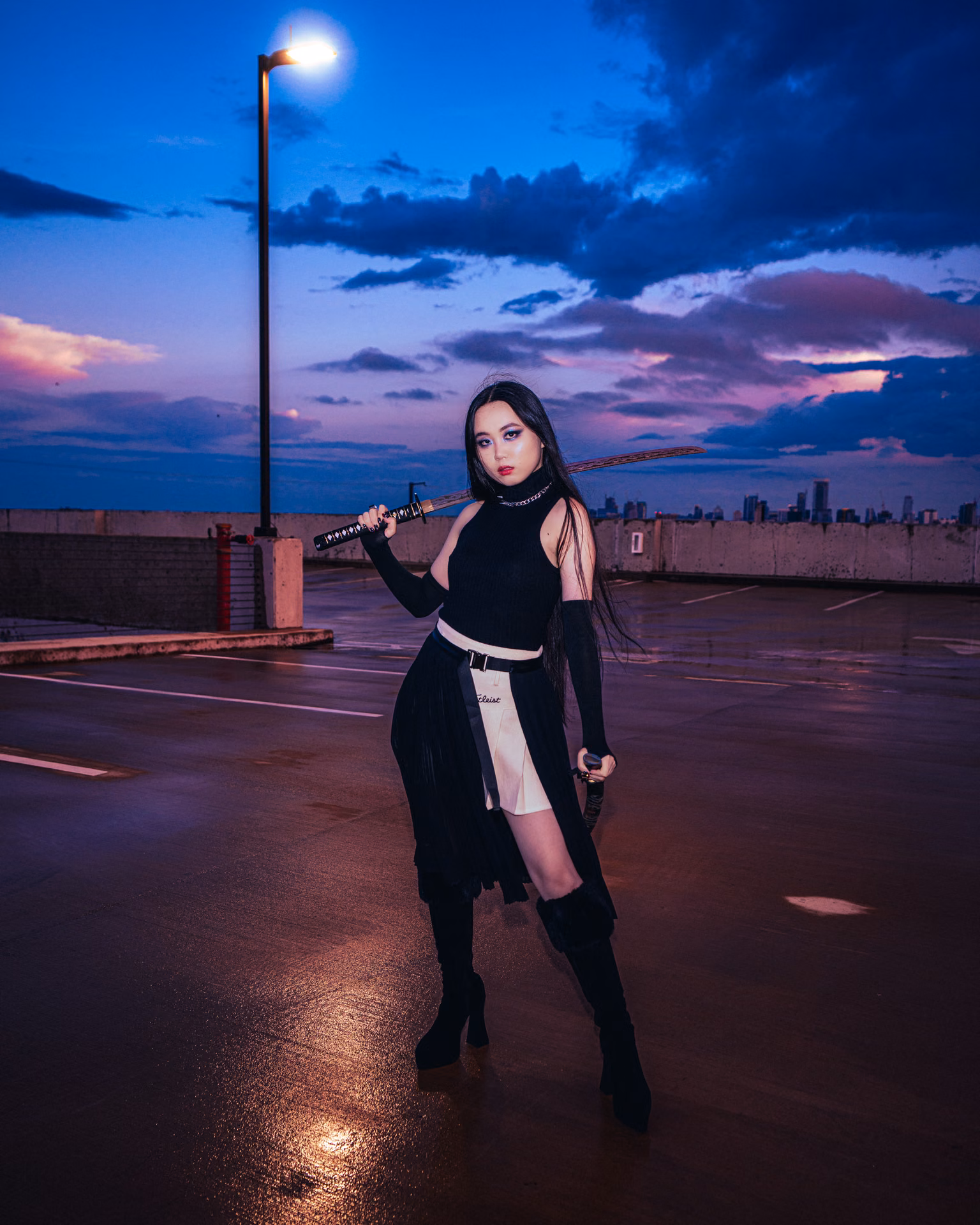At twilight under a pink-and-blue sky, a leather-clad figure rests a blade on one shoulder, moon-and-star chains catching the last light — pure rooftop narrative.