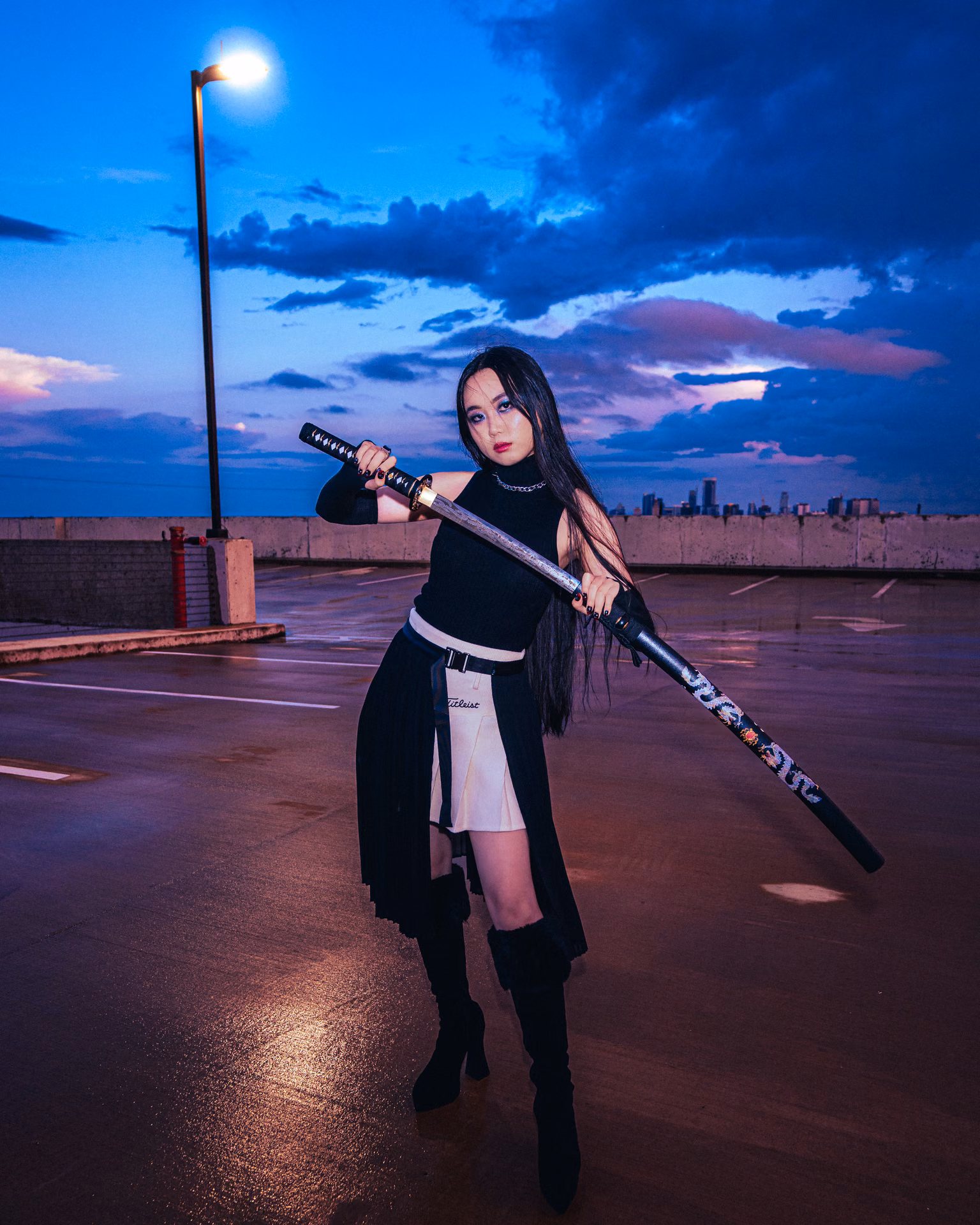 At twilight under a pink-and-blue sky, a leather-clad figure rests a blade on one shoulder, moon-and-star chains catching the last light — pure rooftop narrative.