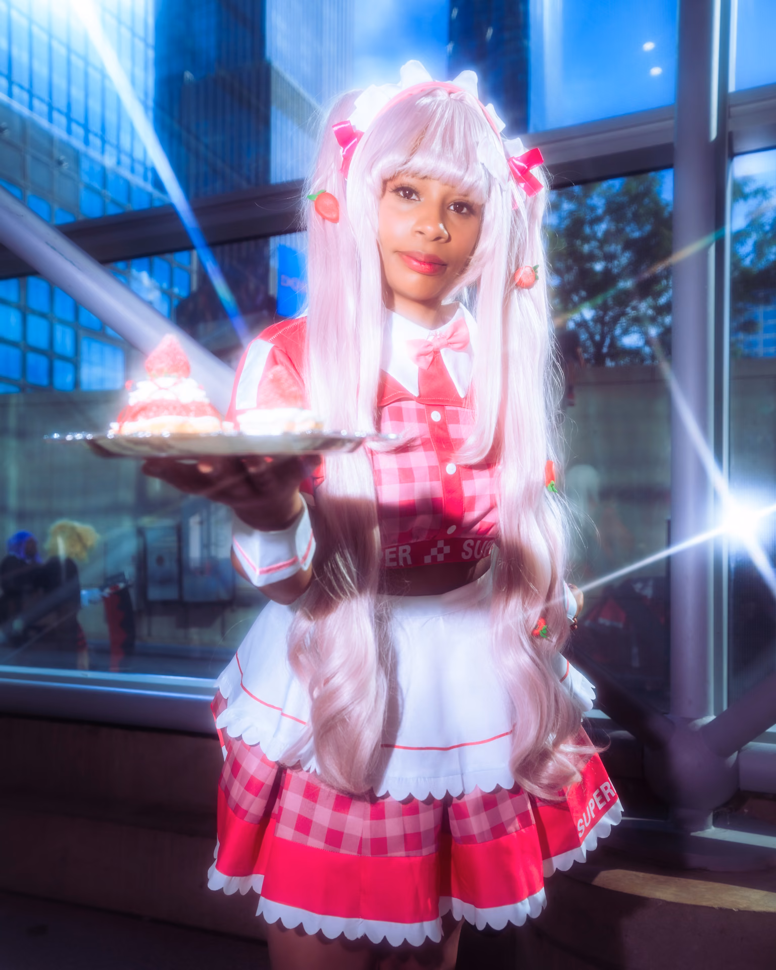 A pink-gingham maid offers a plate of strawberry shortcake through a burst of lens flare, a glass tower behind her; sugar-sweet cosplay against a hard architectural backdrop.